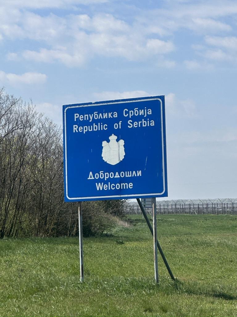 Blaues Grenzschild „Republic of Serbia — Welcome“ an einer grünen Grenze.