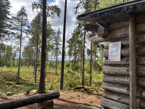 Holzshelter am Waldrand mit Hinweisschild, Blick in lichten Wald.